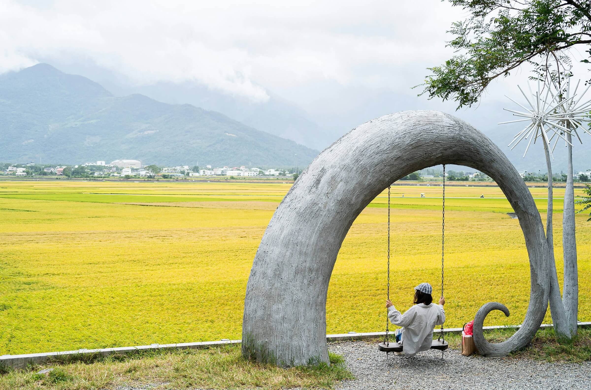 Person sitting on a swing beneath an arched structure in an open landscape.