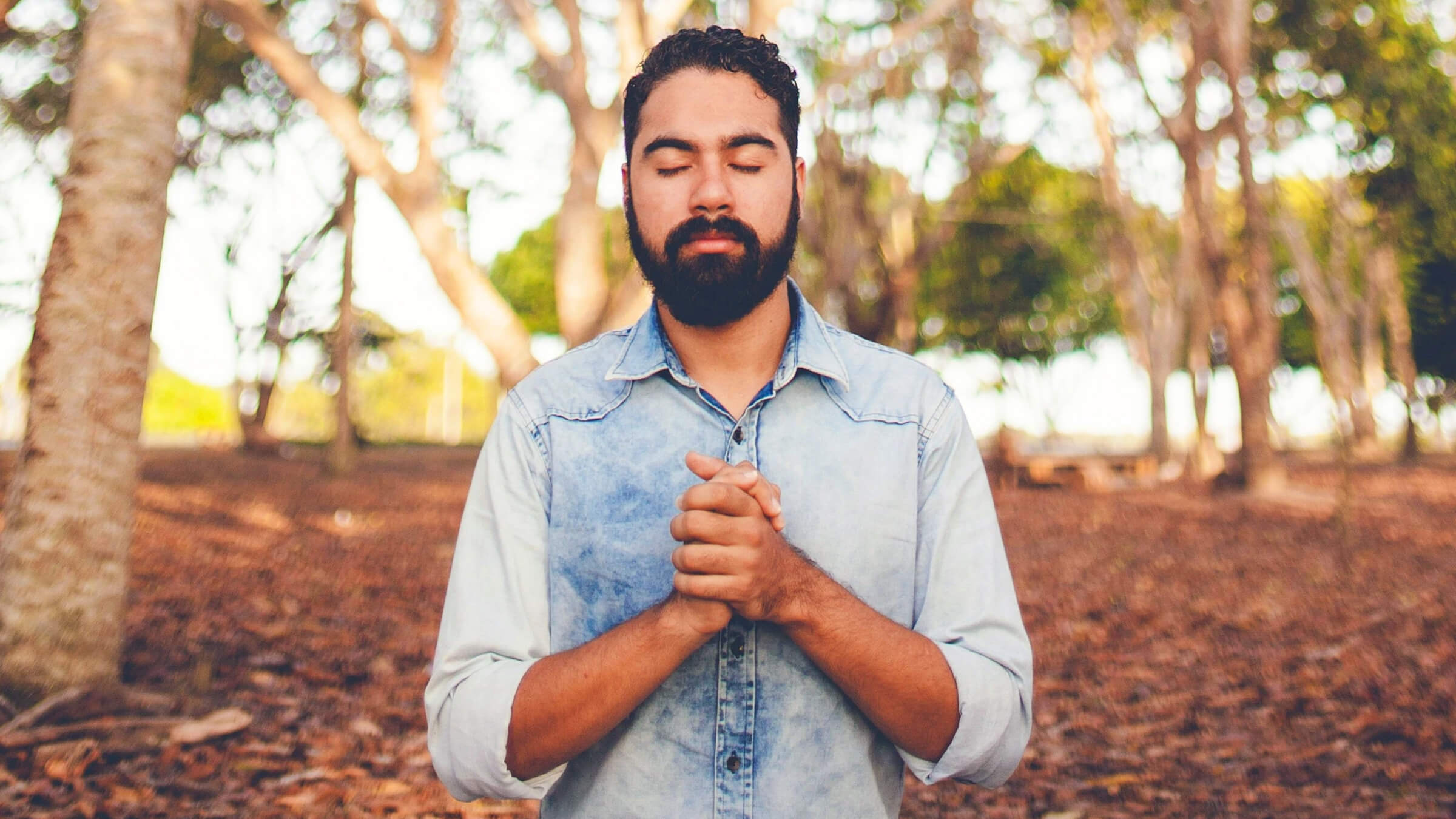 Man standing outdoors with eyes closed and hands clasped, practicing mindfulness meditation among trees